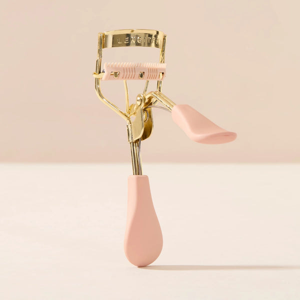 Eyelash Curler Lash Lift by Lenoites, featuring a pink eyelash curler with gold details and a black rubber pad, accompanied by a small pink comb, displayed against a soft beige background.